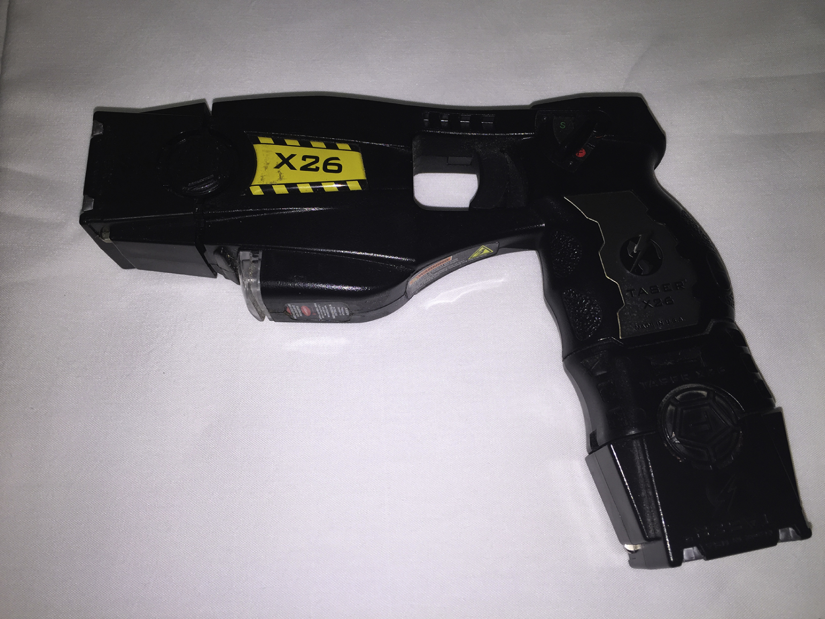 The photograph shows black conducted energy weapon labeled Taser X26. The device has a pistol shape with a textured grip, a trigger, and a yellow label marked “X26” on the upper side.