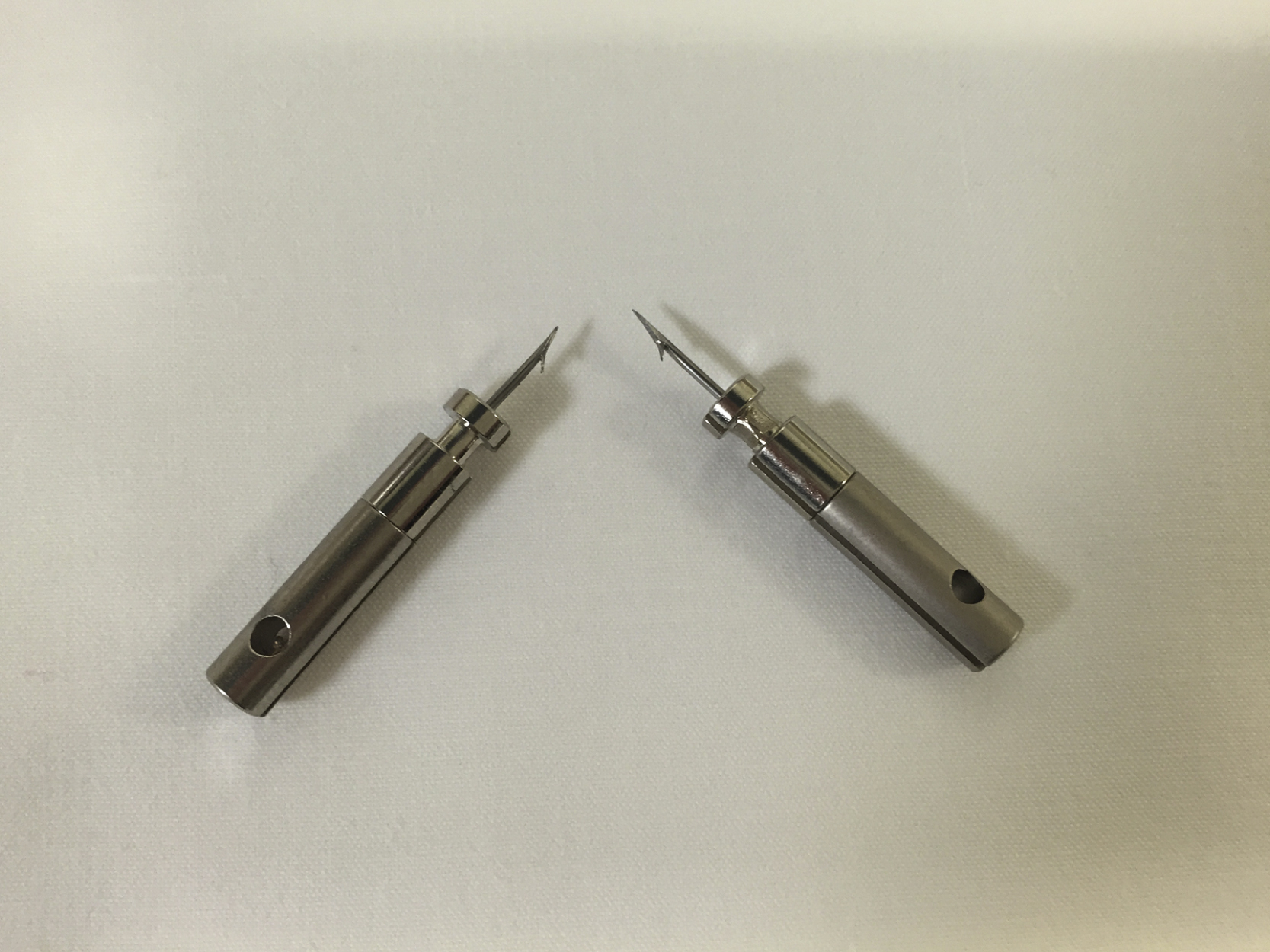 The photograph shows two taser barbs lie on a light surface, each with cylindrical metal body, a hole near one end, and a sharp pointed tip designed to deliver electrical bursts for incapacitation. The photograph shows two taser barbs on a light-colored surface. Each taser barb has a cylindrical metal body with a hole near one end and a sharp pointed tip at the other end designed for penetration. These taser barbs are used to deliver an electrical burst intended to cause neuromuscular incapacitation.
