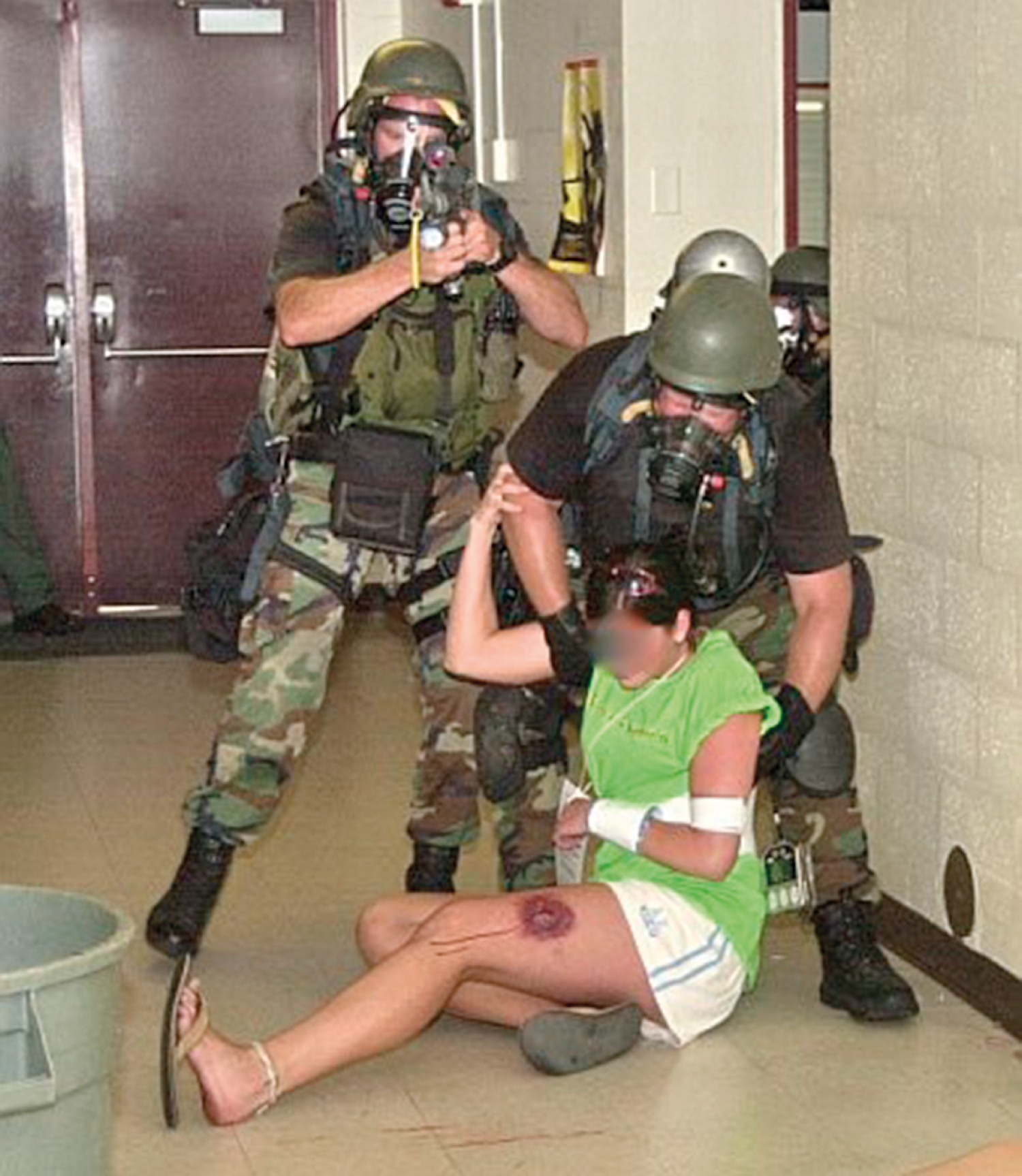 The photograph shows two armed personnel in camouflage and helmets assist an injured woman with a leg wound on the floor, one providing medical aid and the other security cover with a raised rifle. The photograph shows two armed personnel in camouflage uniforms, helmets, and tactical gear assisting an injured woman seated on the floor with a visible leg wound. One rescuer supports and moves the injured woman while another provides security cover with a rifle aimed forward, shows a direct threat phase of care in a high-risk environment such as an active tactical situation or hostile area.