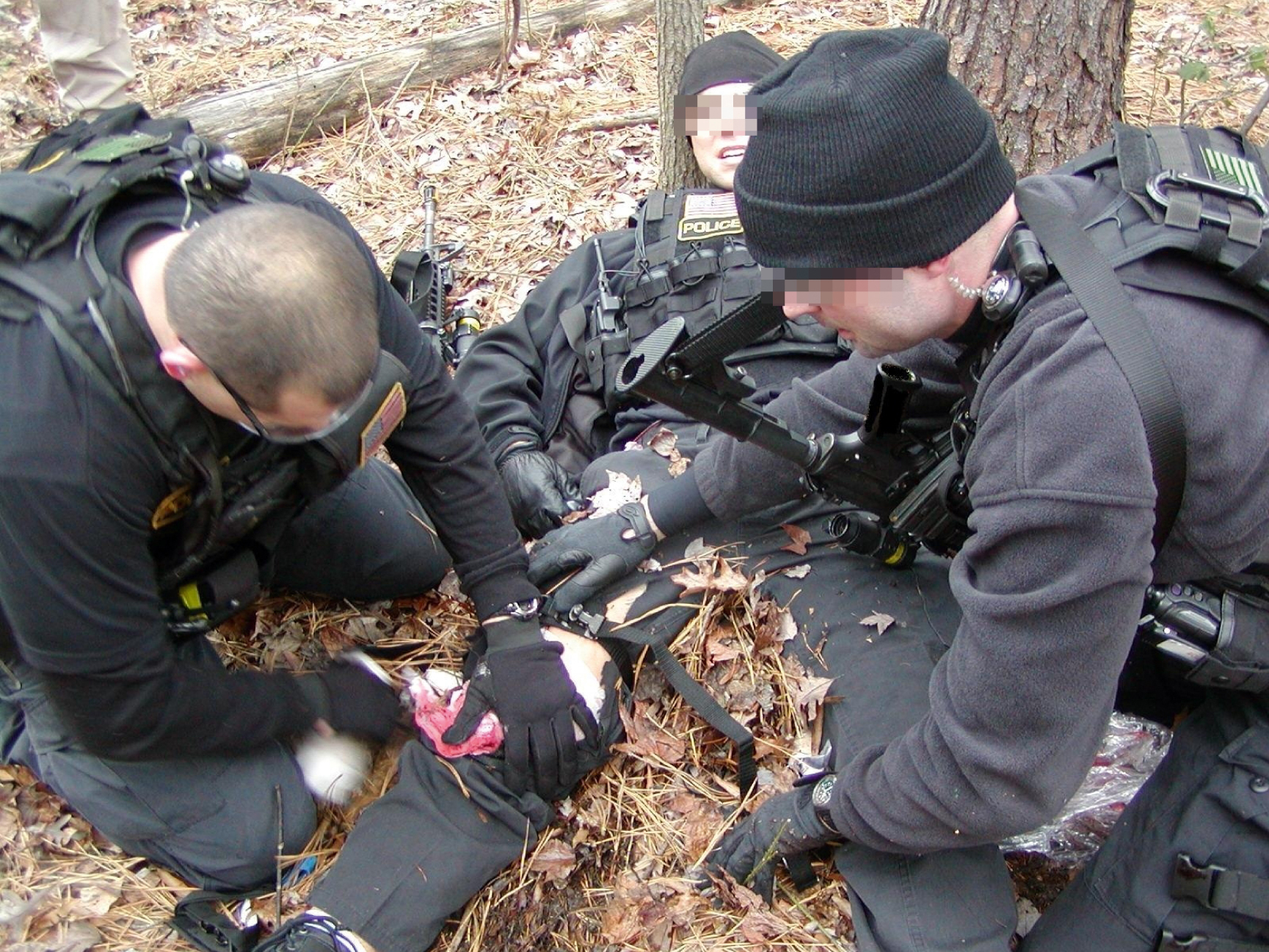 The photograph shows a tactical personnel in black uniforms and gear provide medical aid to an injured person on a leaf covered ground, one applying a leg bandage others assist and secure the area. The photograph shows a tactical personnel in black uniforms and protective gear providing medical aid to an injured individual lying on the ground covered with leaves in a wooded area. One rescuer applies a bandage or dressing to a leg wound while others assist and secure the area, shows tactical field care during an indirect threat phase of care.