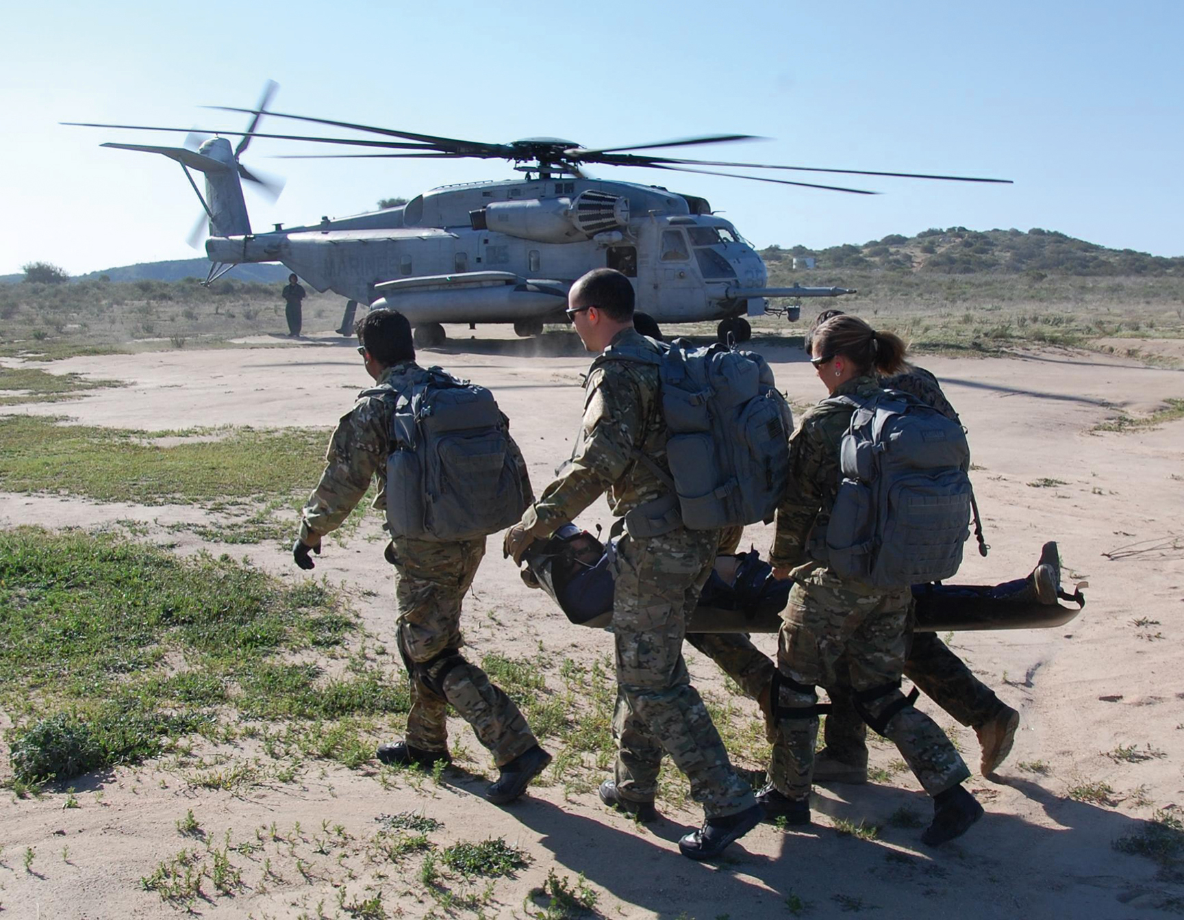 The photograph shows three military personnel carry an injured person on a stretcher toward a running helicopter in an open area, represents combat casualty evacuation care during evacuation phase. The photograph shows three uniformed military personnel carrying an injured individual on a stretcher toward a waiting military helicopter on a flat open area, shows combat casualty evacuation care during the evacuation phase. The helicopter has its rear door open and rotors spinning, ready for rapid transport from the field.